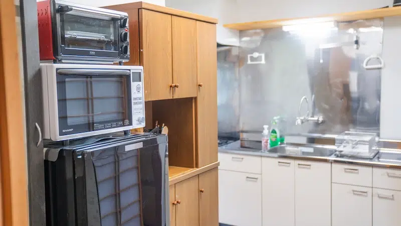 Kitchen corner with a black refrigerator, white microwave, and a red toaster oven stacked on top, next to wooden cabinets.