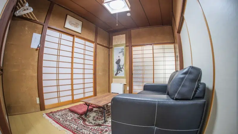 Cozy Japanese-style room with sliding shoji doors, low wooden table, red cushion, and two dark leather armchairs on a patterned rug.